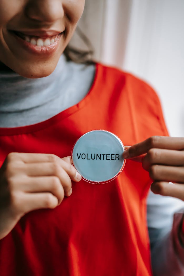 Crop anonymous positive ethnic female smiling and demonstrating sign with inscription volunteer on bright red apron